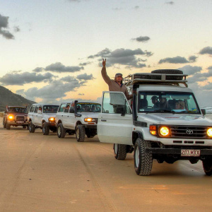 Convoy of 4WD vehicles driving along the beach at sunset on K'gari Fraser Island, with a tour guide celebrating through the sunroof