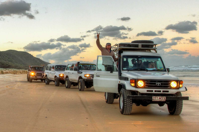 Convoy of 4WD vehicles driving along the beach at sunset on K'gari Fraser Island, with a tour guide celebrating through the sunroof