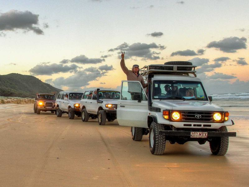Convoy of 4WD vehicles driving along the beach at sunset on K'gari Fraser Island, with a tour guide celebrating through the sunroof